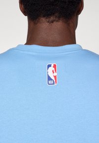Svetlomodré bavlnené tričko s okrúhlym výstrihom, so bielym nášivkou loga NBA na zadnej strane. Hladká textúra a jednoduchý dizajn.