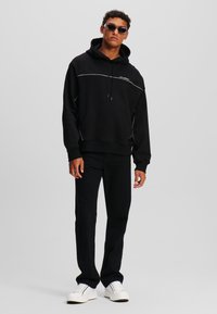 Haut à capuche noir avec une poche kangourou, détails de coutures blanches, associé à un pantalon noir et des baskets blanches avec des accents bleus.