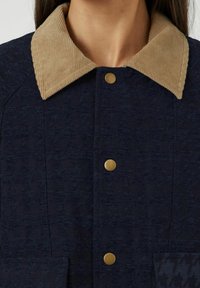 Marinblå tygjacka med ett strukturerat mönster, utrustad med en beige kordflorskrage, gyllene tryckknappar och en framficka.