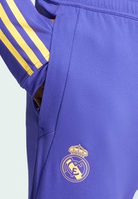 Purple athletic pants featuring yellow stripes on the sides, a zippered pocket, and a gold embroidered club logo on the lower leg.