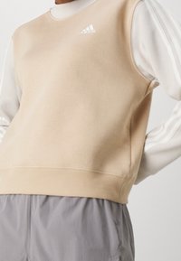 Béžová sleeveless mikina s bílými dlouhými rukávy, žebrovaným lemem a logem Adidas na hrudi; měkký bavlněný materiál; volný střih.