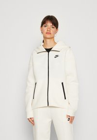 Balta vilnonė striukė su priekine užtrauktuko kišene, juodais akcentais ir gobtuvu. Ant krūtinės yra mažas juodas Nike logotipas ir užtrauktuku užsegamos kišenės.