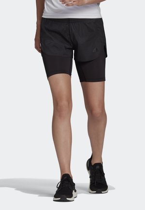 Person trägt schwarze mehrlagige Laufshorts, schwarze Laufschuhe und ein weißes Oberteil, steht vor hellem Hintergrund.