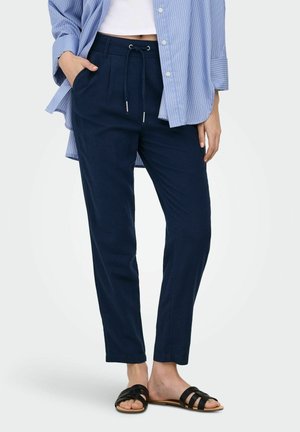 Femme portant un pantalon à cordon de serrage bleu marine, un haut blanc court, une chemise rayée bleue et des sandales noires à enfiler, debout devant un fond uni.