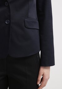 Detalle de una persona que lleva un blazer azul marino oscuro con botones y un bolsillo con solapa, combinado con pantalones negros.
