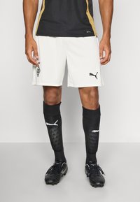 Bela nogometna kratka hlača z črnim logotipom in značko Puma, v kombinaciji s črnimi nogavicami z belimi detajli in teksturiranimi paneli. Črni čevlji.