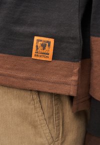 Πουκάμισο με μακριά μανίκια σε σκούρο γκρι και σκουριασμένο χρώμα, με ραμμένη πορτοκαλί ετικέτα Brixton, συνδυασμένο με ριμπ ύφους μπεζ βελούδινο παντελόνι.