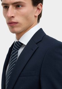 Navy(coloured) jasje met notch-lapel, voorzien van een borstzak aan de linkerzijde en een getailleerde pasvorm. Gedragen met een lichtblauw overhemd met kraag en een gestreepte das.