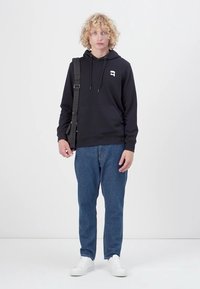 Hanorac negru cu buzunare laterale și logo pe piept, asortat cu blugi din denim albastri și adidași albi. Design simplu, cu șnururi.