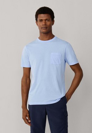 Man staat met hand in zak, draagt een lichtblauw T-shirt met korte mouwen en borstzak, en donkerblauwe cargobroek tegen een effen achtergrond.