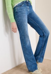 Personne portant un jean bicolore bleu, un pull rayé vert clair et des baskets blanc cassé, debout contre un mur blanc et beige.