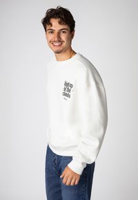 Weißes Sweatshirt mit Rundhalsausschnitt und gerippten Bündchen. Es hat einen grünen Druck mit dem Schriftzug "hoch in den Wolken". Getragen mit blauen Jeans.