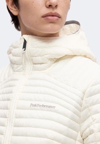 Personne portant une veste matelassée crème clair avec capuche, fermeture éclair devant et logo Peak Performance brodé sur la poitrine.