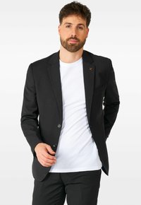 Blazer negro con dos botones, solapas de pico y un bolsillo en el pecho, llevado sobre una camiseta blanca, combinado con pantalones negros.