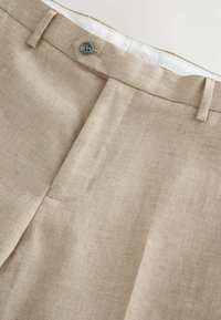 Pantalones de lino beige con un acabado texturizado, que presentan un cierre de un solo botón, una cintura bien cosida y un diseño con pliegues en la parte delantera.