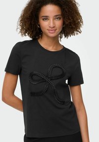 Ung kvinde med krøllet hår iført en sort kortærmet skjorte med et tekstureret sløjfedesign på fronten, der står foran en hvid baggrund.