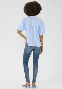 Blusa azzurro chiaro a maniche corte con maniche a sbuffo, cucitura a yoke sul retro e vestibilità comoda, abbinata a jeans blu attillati e scarpe slip-on grigie.