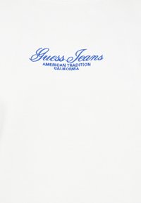 Witte T-shirt met korte mouwen en blauwe geborduurde tekst. Bevat "Guess Jeans" en "American Tradition California" in cursieve stijl.