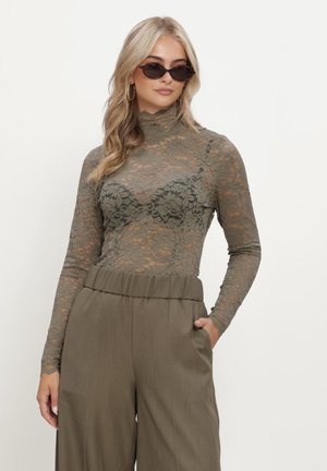 Woman wearing olive lace long-sleeve top and matching high-waist wide-leg pants, with dark sunglasses and gold hoop earrings, hand in pocket.