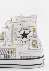 Białe trampki Converse Chuck Taylor z wysoką cholewką, z kolorowym nadrukiem kaset magnetofonowych i czarną naszywką z logo gwiazdy na kostce.