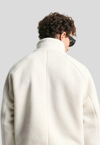 Fleece jakna u boji bijele kave, s visokim ovratnikom i teksturiranim materijalom. Dizajana je s opuštenim krojem i kontrastnim šavovima.