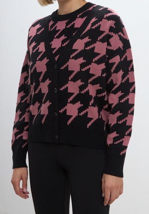 Femme portant un cardigan boutonné avec un grand motif pied-de-poule rose et noir, associé à un pantalon noir, visible des épaules aux hanches.