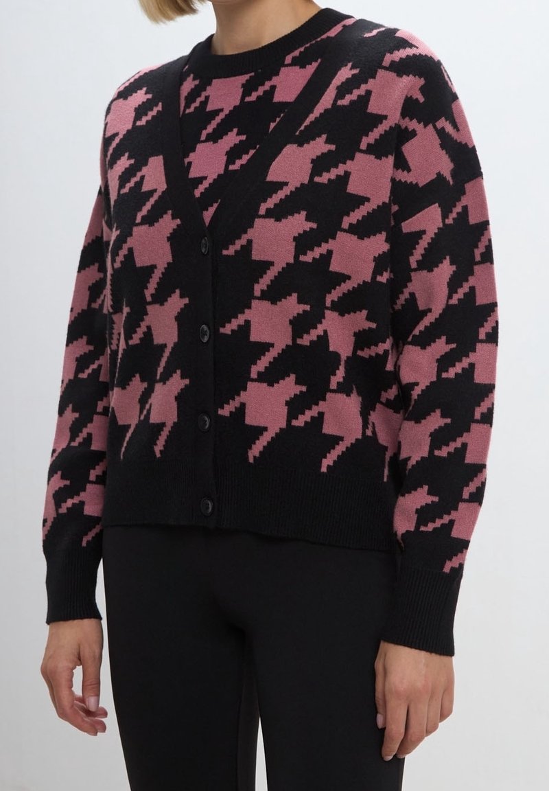 Femme portant un cardigan boutonné avec un grand motif pied-de-poule rose et noir, associé à un pantalon noir, visible des épaules aux hanches.