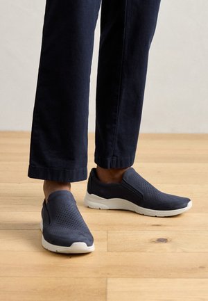 Scarpe slip-on grigio scuro con tomaia perforata, suola bianca e design senza cuciture. Indossate con pantaloni cropped blu navy su un pavimento di legno.