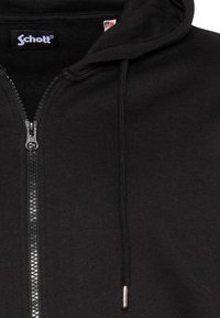 Sudadera con capucha negra con cremallera, confeccionada en un tejido suave, presenta una cremallera metálica, cordones ajustables y una etiqueta con un detalle de la bandera estadounidense.