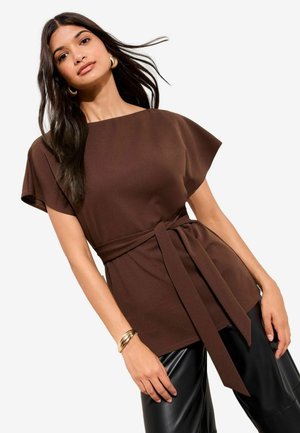 Frau mit langen dunklen Haaren, die goldene Creolen, ein braunes, kurzärmeliges, mit Gürtel versehenes Oberteil, schwarze Lederhosen und ein goldenes Armband trägt.