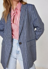 Blazer bleu à rayures en tissu léger, avec une fermeture à un bouton, des revers en notch et deux poches avant sur une chemise à rayures rouges.