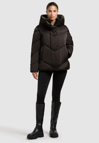 Giacca a vento nera con colletto alto, con un motivo a zigzag e chiusura con zip. Indossata con leggins neri e stivali neri fino al ginocchio.