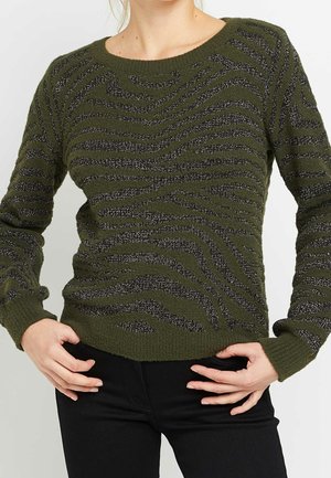 Femme portant un pull en maille vert olive avec des rayures zébrées noires scintillantes et un pantalon noir, les mains sur les hanches.