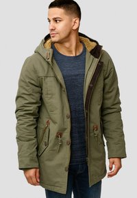 Olivenfarget parka med hette, med faux pelsfôr, flere lommer og brune detaljer. Myk, strukturert stoff med en avslappet passform.