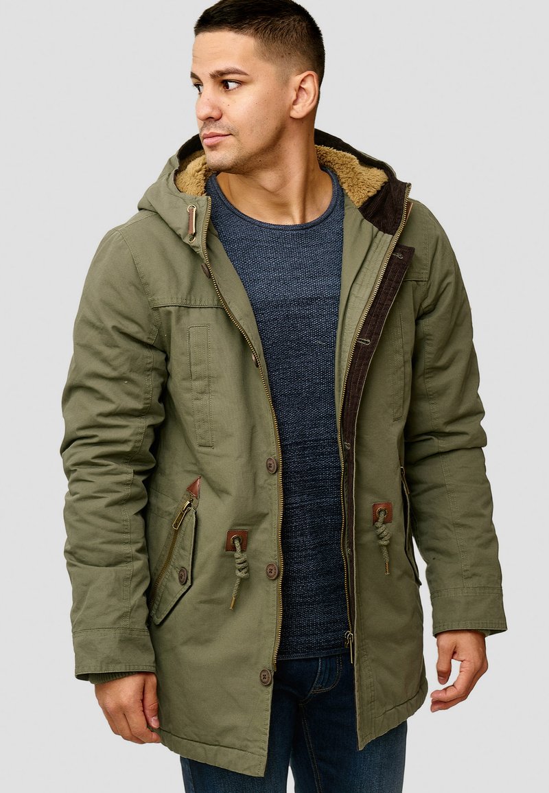 Olivenfarget parka med hette, med faux pelsfôr, flere lommer og brune detaljer. Myk, strukturert stoff med en avslappet passform.