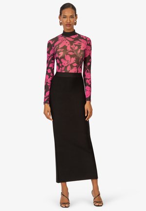Femme portant un haut ajusté à manches longues, rose et noir à motifs floraux, rentré dans une jupe noire taille haute et longueur cheville, accompagnée de talons noirs à lanières.