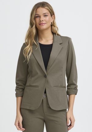 Frau trägt olivgrünen Blazer mit hochgekrempelten Ärmeln über schwarzem Oberteil, passende Hose und goldene Creolen vor schlichtem weißen Hintergrund.