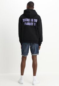 Hombre llevando una sudadera con capucha negra con el texto "NO HAY UN PLANETA B" en la espalda, pantalones cortos de mezclilla, calcetines blancos y zapatillas blancas, de pie mirando hacia atrás.