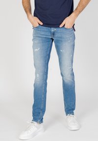 Vaqueros de mezclilla azul claro con detalles desgastados, corte slim, cinco bolsillos y cremallera, combinados con zapatillas blancas.