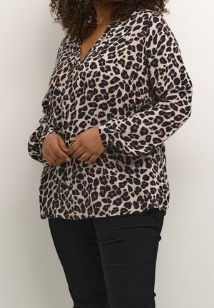 Woman with curly hair wearing a long-sleeve beige blouse with black leopard print and black pants, standing against a plain background.