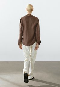 Brun, långärmad skjorta med en slät textur, kombinerad med ljusa, mönstrade vida byxor. Modellen ses bakifrån.