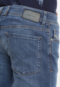 Vaqueros de mezclilla azul con acabado desgastado, con parche de cuero de la marca DIESEL, bolsillos traseros estándar y detalles de costura.