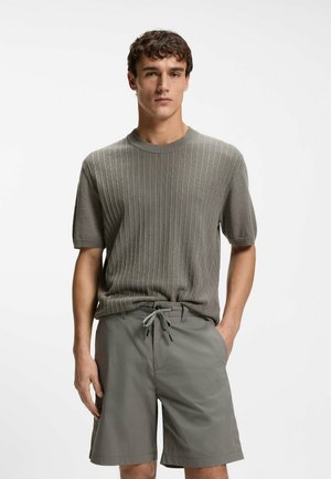 Mand iført en grå ribstrikket kortærmet sweater og grå shorts med snøre, stående med den ene hånd i lommen foran en ensfarvet baggrund.