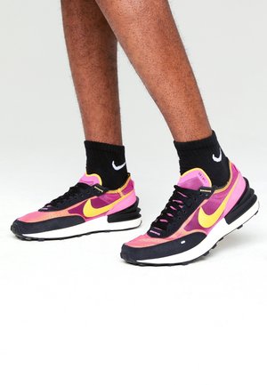Personne portant des baskets Nike roses et noires avec des virgules jaunes et des chaussettes Nike noires, debout sur un fond blanc.