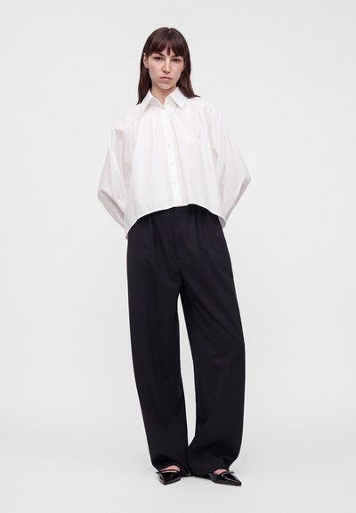 Femme aux cheveux bruns raides porte une chemise blanche ample boutonnée, un pantalon large noir et des chaussures noires pointues, debout devant un fond uni.