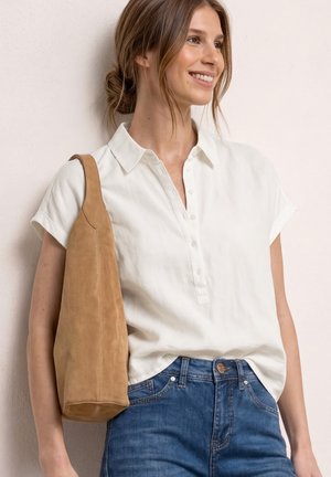 Giovane donna con camicia bianca a maniche corte abbottonata e jeans blu porta una grande borsa a tracolla in camoscio marrone, sorridente e guardando a destra.