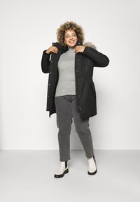 Parka noire avec capuche en fausse fourrure, taille cintrée et poches latérales, associé à un pull gris en côtes et un jean gris, bottines noires et blanches.