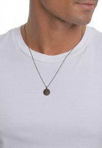 Bronzas kulons uz plānas melnas ķēdītes ar teksturētu apaļu dizainu, nēsāts virs vienkāršas balti T-krekla.