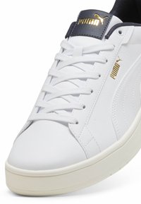 Hvide sneakers lavet af syntetisk læder med en tekstureret overflade, der har hvide snørebånd, en mørkeblå tunge og gyldne branddetaljer.