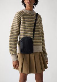 Schwarze Umhängetasche mit abgerundeter Form, aus Nylonstoff gefertigt, mit einem dezent geprägten Logo und verstellbarem Tragegurt, getragen mit einem gestreiften Pullover.